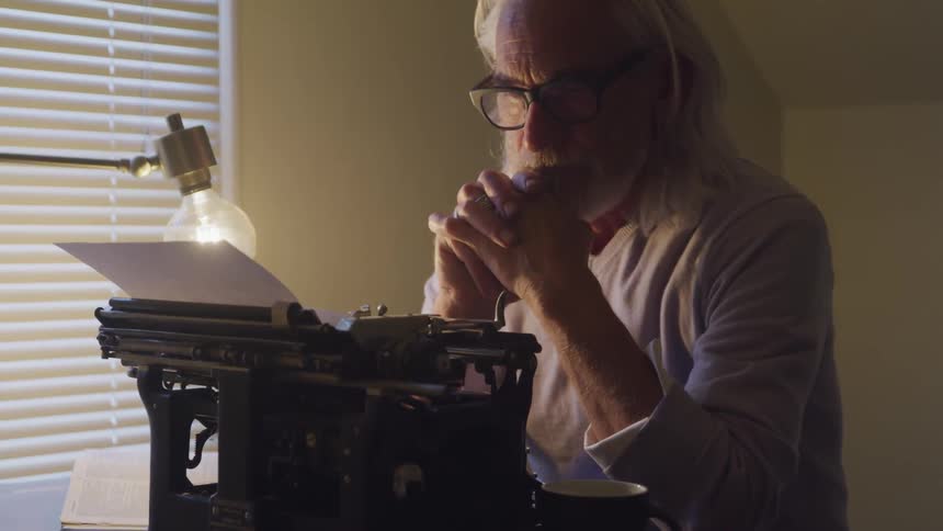 Man sitting at the table looks at the typewriter and reads what he has written in a room