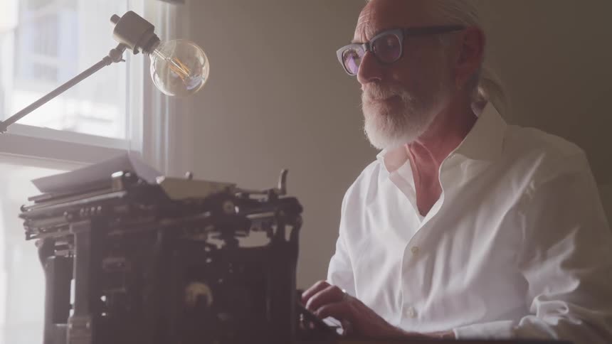 Older man typing on a vintage typewriter near a window