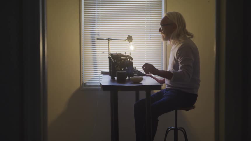 An older man with longish white hair sits at a small table with a mechanical typewriter and sets down his cigarette
