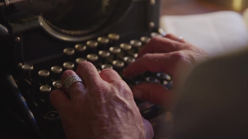 Senior man sitting at the table types on typewriter in a room