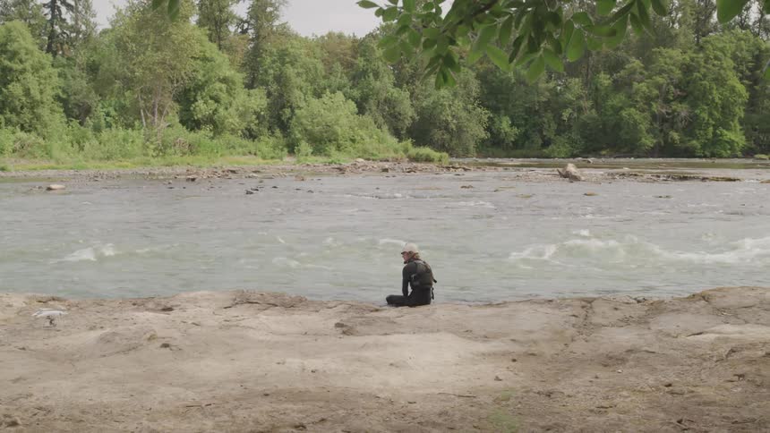 A person sits on a dirt bank beside a fast moving river