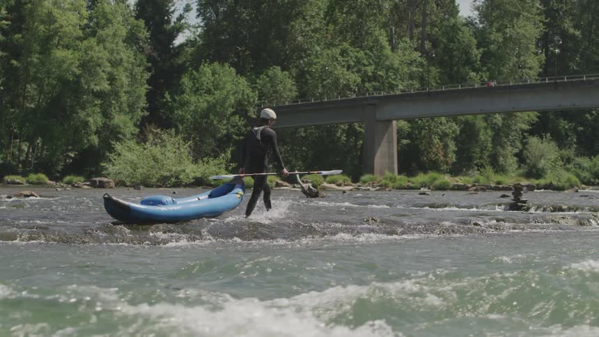 A man in a full body wet suit is dragging a kayak through shallow rapids in a river near a bridge while carrying the paddle in his other hand