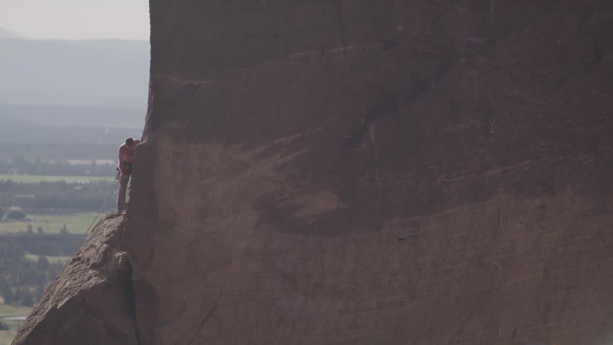 A male rock climber stands on a small ridge and pulls up a long rope hanging below him