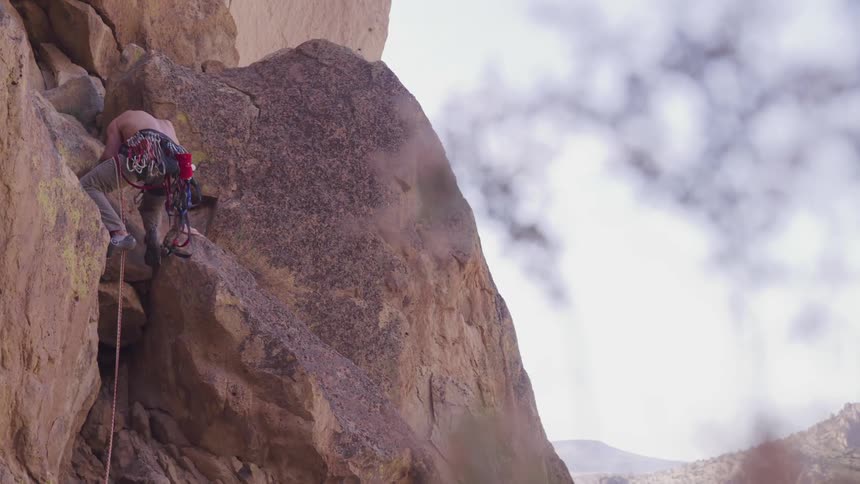 A shirtless white male equipped with climbing gear is ascending a steep rock face with mountains in the distance
