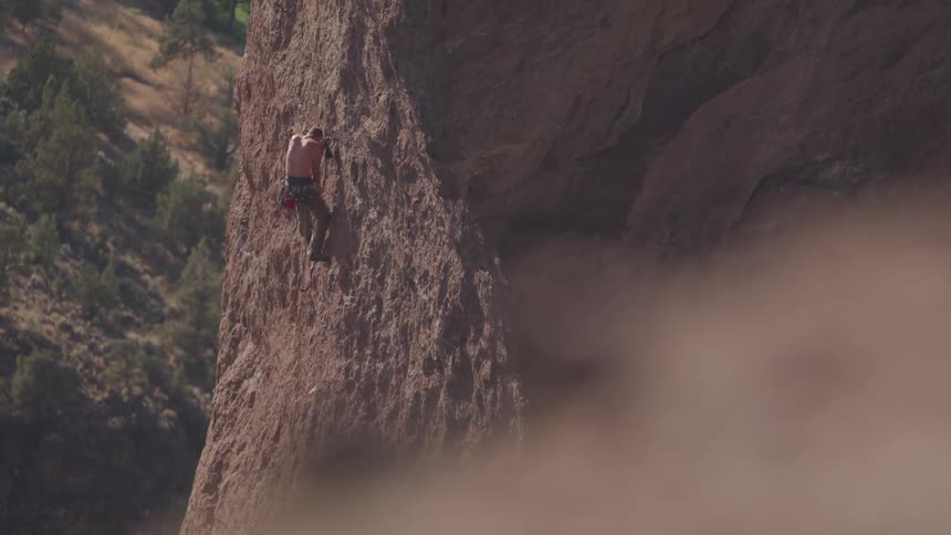 A shirtless rock climber makes his way up the side of a cliff