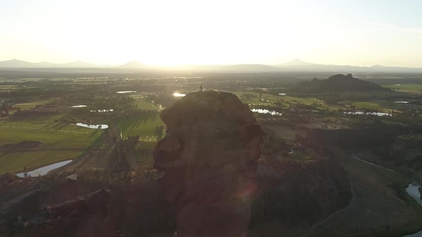 A man stands on top of a mountain peak in a rural area