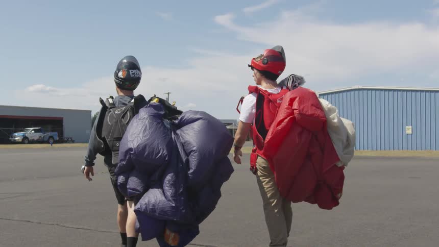 Two men carrying holding deployed parachutes across the tarmac