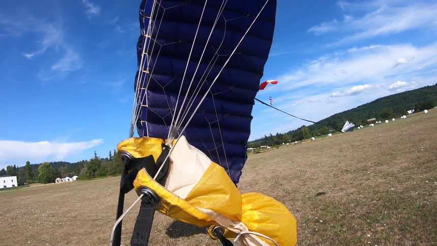 A wide open grass field can be seen from the helmet mounted camera of a person who is parachuting to the ground