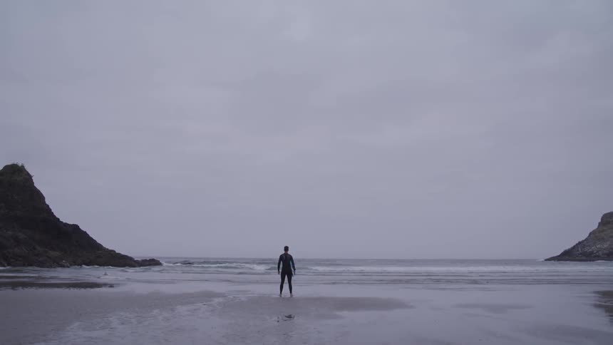 A man in a wet suit stands on the beach while holding his surfboard on an overcast day
