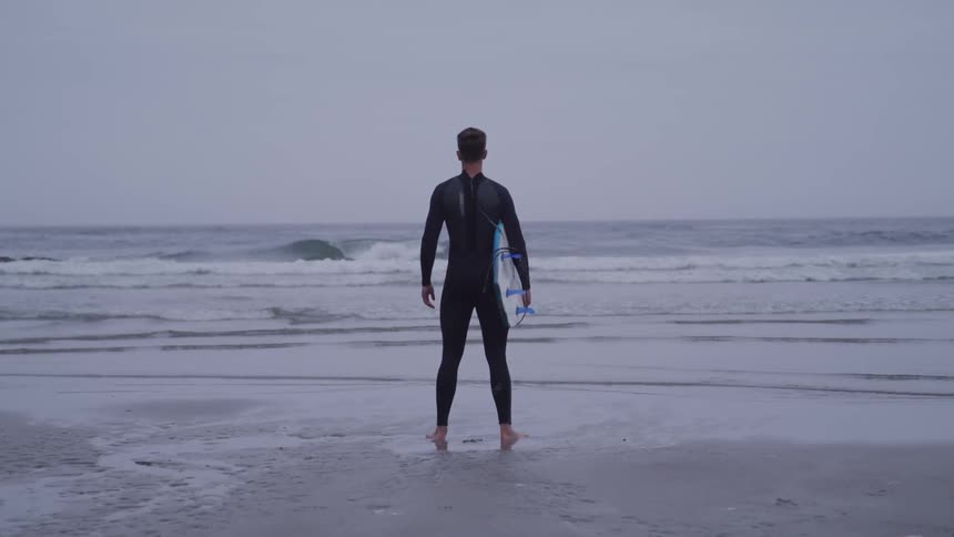 A man holding a surfboard and wearing a wet suit stands looking out at the ocean waves