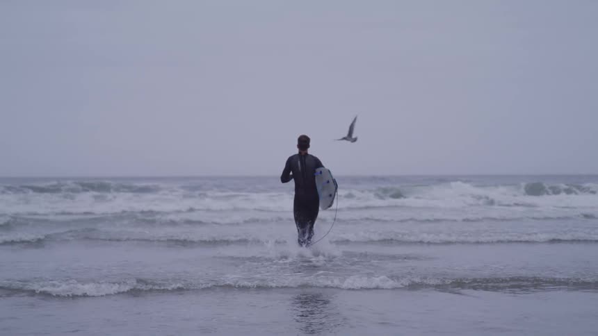 Man in wetsuit holding surfboard goes swimming on cloudy day