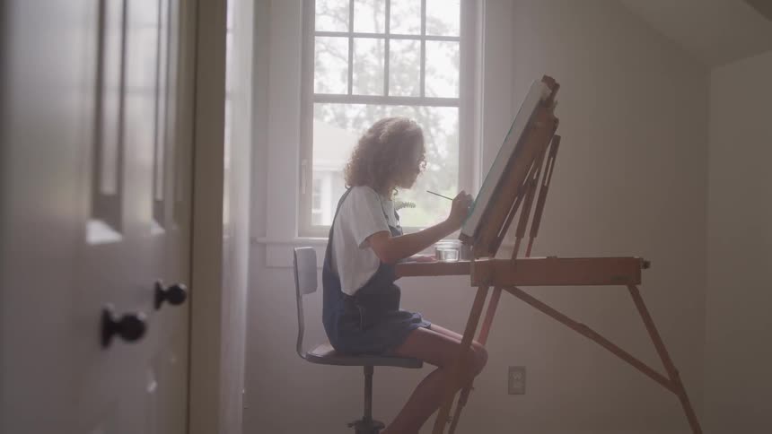 Young girl sitting next to an attic window painting on a canvas that is propped up on an easel