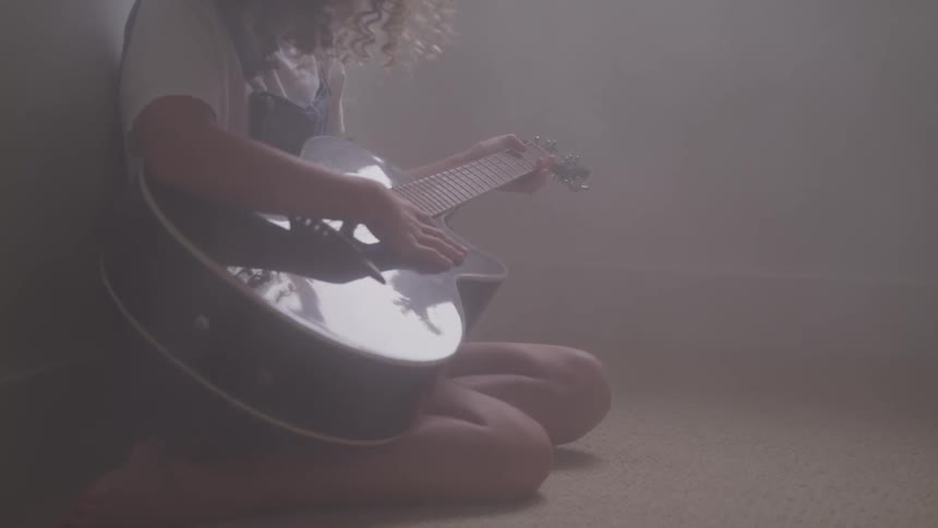 A young female with curly hair sits in a darkened room while strumming an acoustic guitar