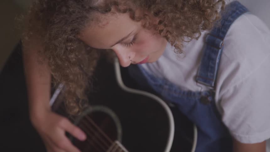 Woman with curly hair wearing overalls playing an acoustic guitar