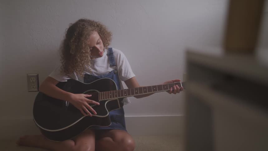 Curly red hair girl plays a guitar leaning against a wall in a room