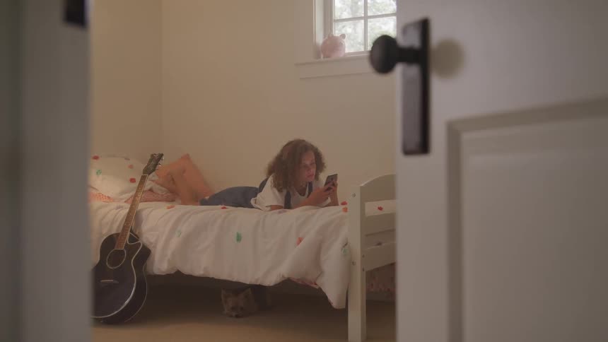 Girl lays on a bed using a cellphone while a guitar sits nearby