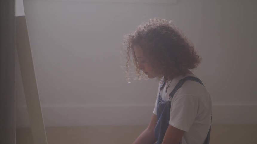 A child with curly hair sits in a room on the floor looking down with head bowed