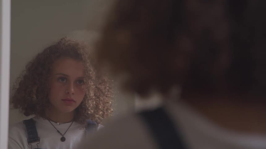A young female with very curly hair looks at her reflection in a mirror indoors