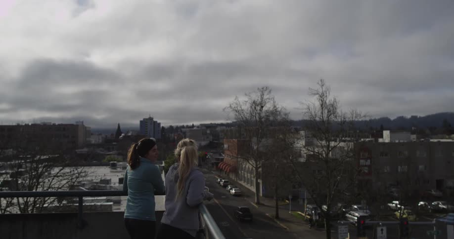Two women stand on a rooftop overlooking a city