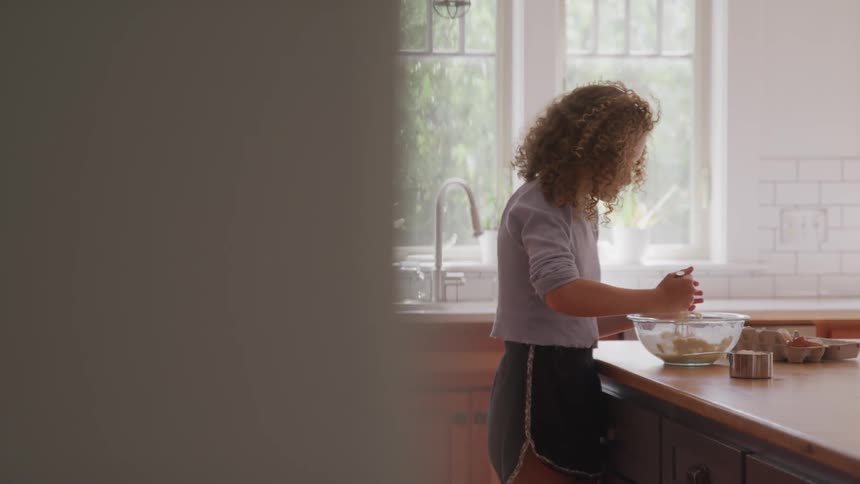 A teenage girl is standing at a kitchen counter stirring a mixture in a glass bowl using a whisk