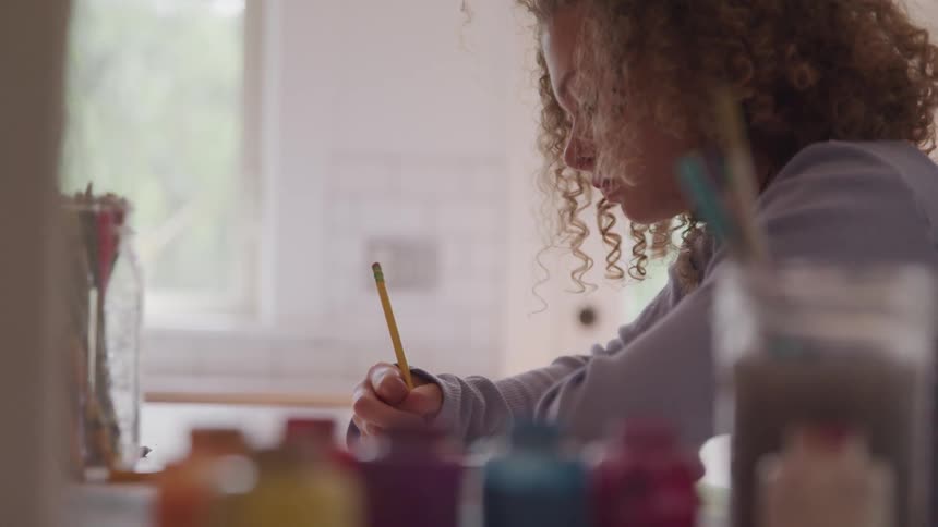 A girl uses a pencil while at a table of art supplies