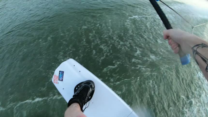 A man with tattoos on his arms holds a towline and does tricks on a wakeboard while being pulled behind a boat on a lake