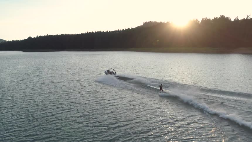 A person being towed behind a boat on a wakeboard moves to ride the wake on an otherwise calm lake