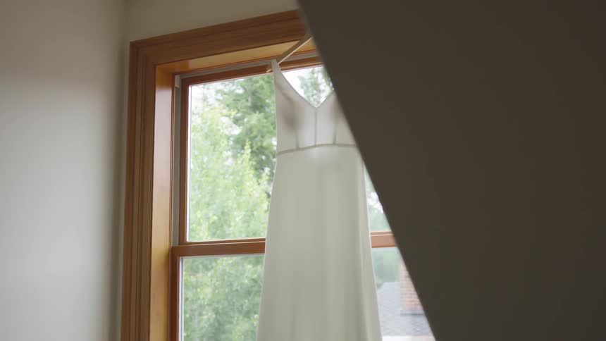 A wedding gown hangs on a hanger in front of a window