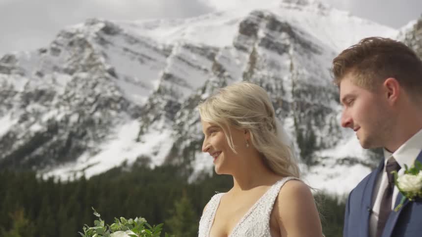A woman wearing a formal gown while holding a bouquet standing next to a man in suit at the base of a snowy mountain