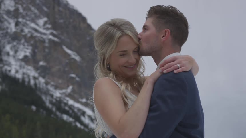 A bride and groom stand outside and hug while the man kisses her