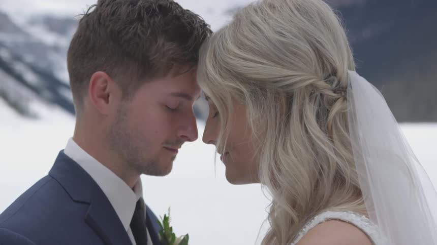 A bride and groom are standing with their eyes closed and foreheads touching with snow and mountains in the background