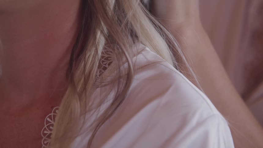 A woman with blonde hair is sitting while wearing a silky robe as someone else styles her hair