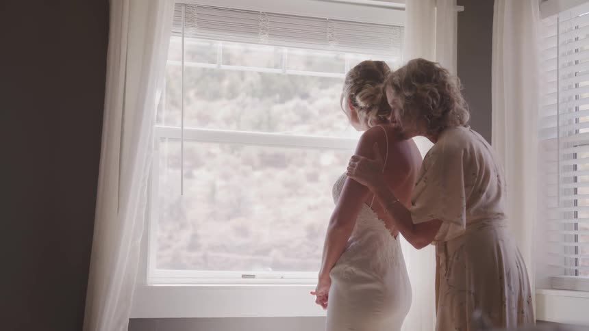 Woman leans in to kiss a bride on her shoulder as the bride looks out a window