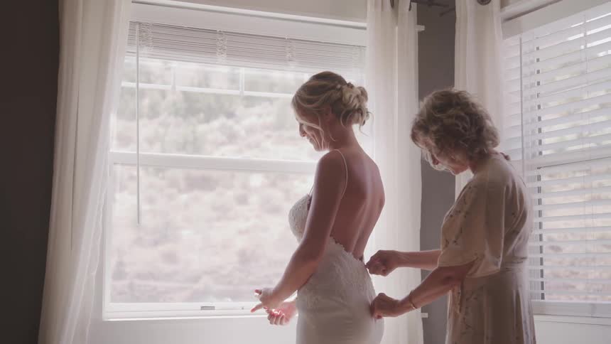 A woman zips up the back of another womans wedding gown