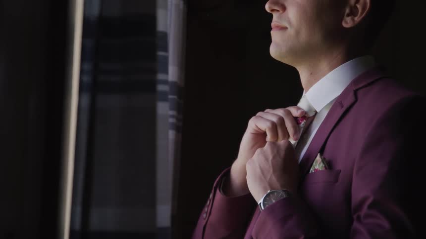 Groom in suit straightens the tie and looks out the window in a room