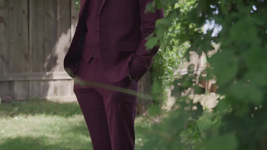 Man in a dark brown suit stands near a bush on a sunny day
