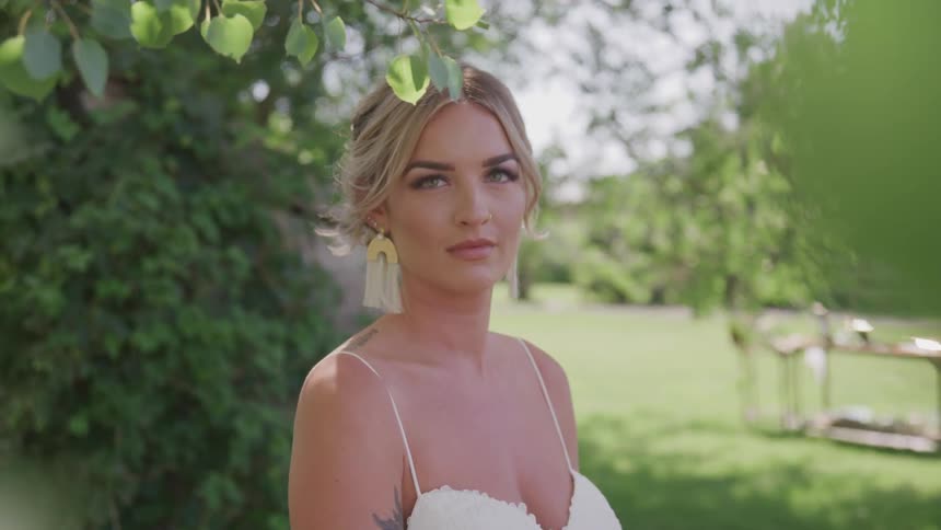 A blonde haired woman in a wedding dress stands under a tree on a sunny day