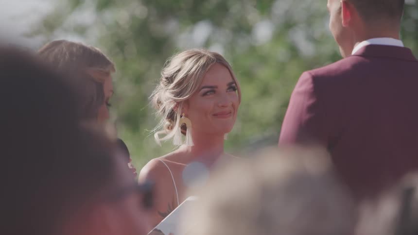 A woman in a wedding dress looks up at her male partner and smiles as another woman speaks into a microphone