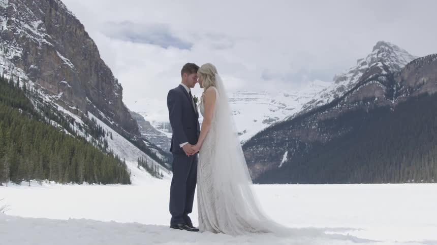 A female in a wedding gown and male in a tuxedo are having wedding photos taken standing outside in the snow with mountains and evergreens in the background