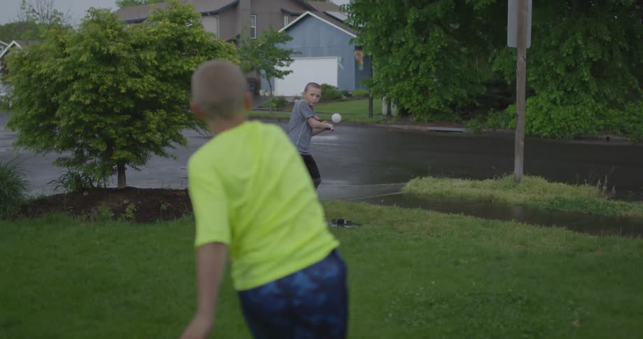 Two young boys play baseball in a grassy yard in a residential area