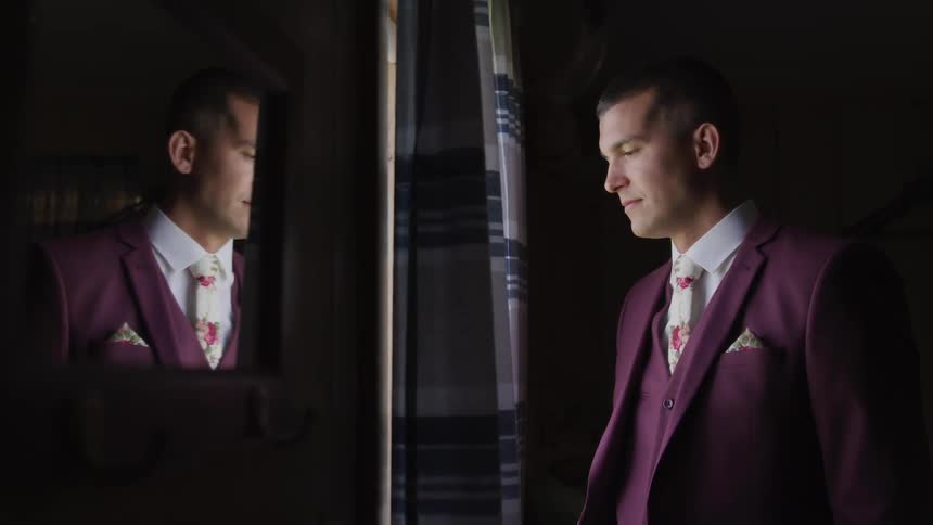 Man in a suit stands and looks out the window in a dark room