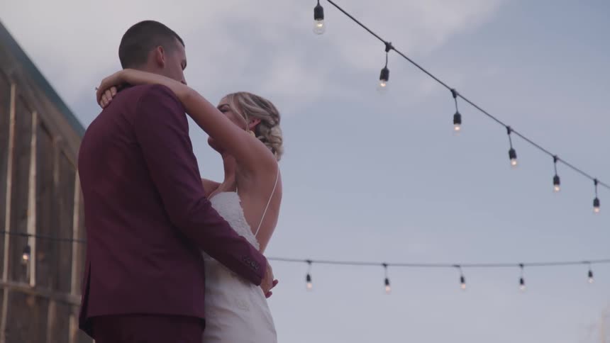 A man in a dark purple suit and a female in a white wedding gown are slow dancing beneath white string lights on a slightly cloudy day