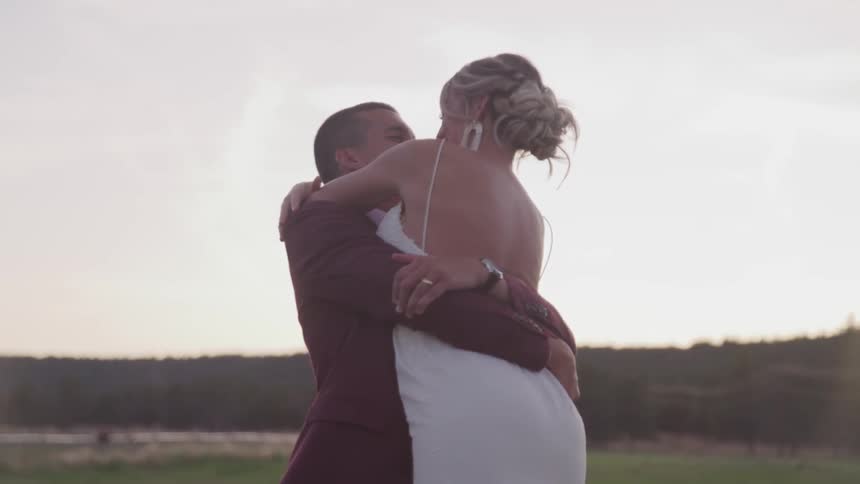 A man in a tuxedo is holding a female in a white dress up in his arms and swinging her around with rollinghills in the background