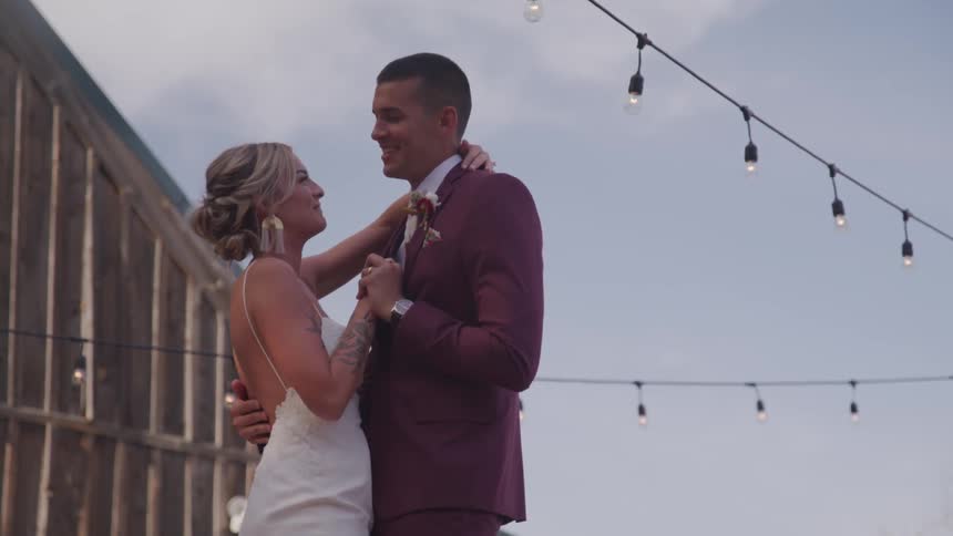 A female in a white wedding gown and man in a deep purple tuxedo are dancing and laughing outside beneath string lights