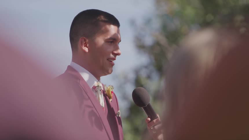 A young man in a gray suit stands outside while talking into a microphone in front of other people