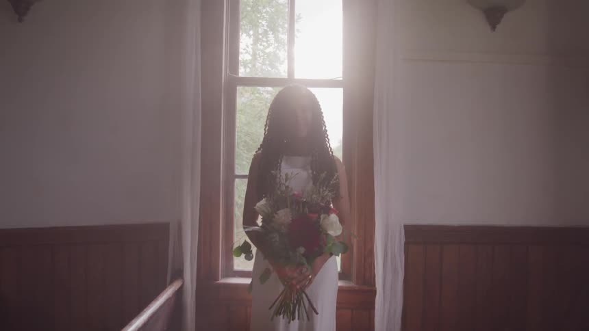 Black bride holding a bunch of flowers stands in front of the church window during wedding ceremony