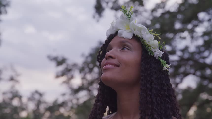 A smiling black woman looks upwards and smiles as she wears a crown of flowers atop her head