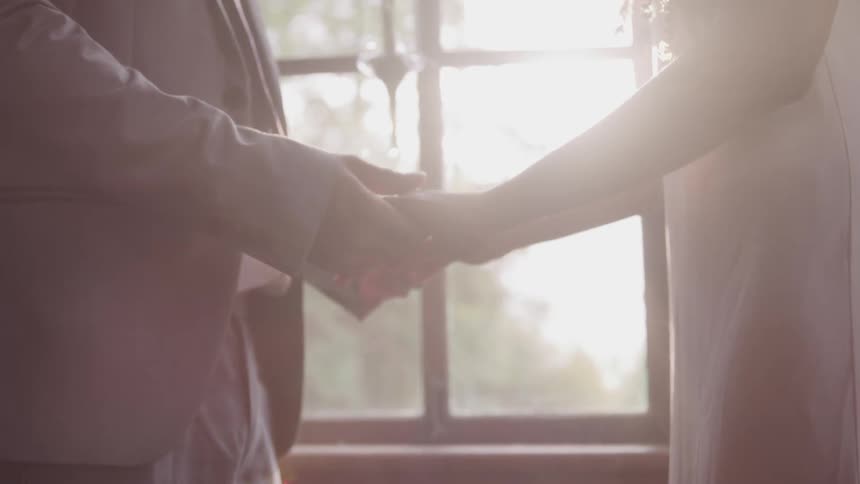 Groom and bride hold hands and look at each other while the sun streams through the window