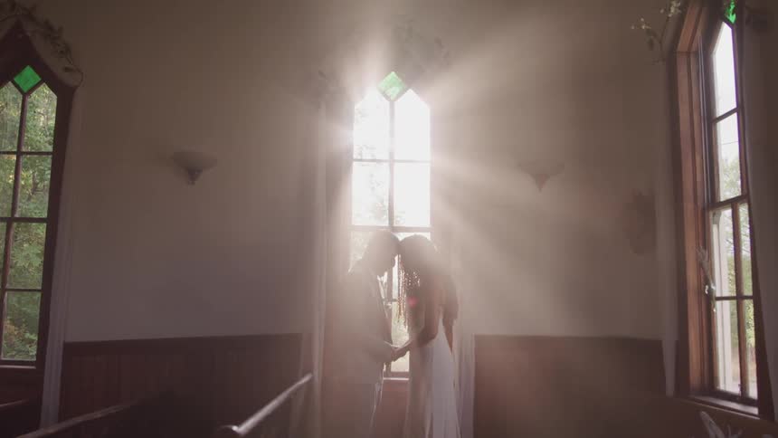 Groom and bride hold hands and look at each other while the sun streams through the window