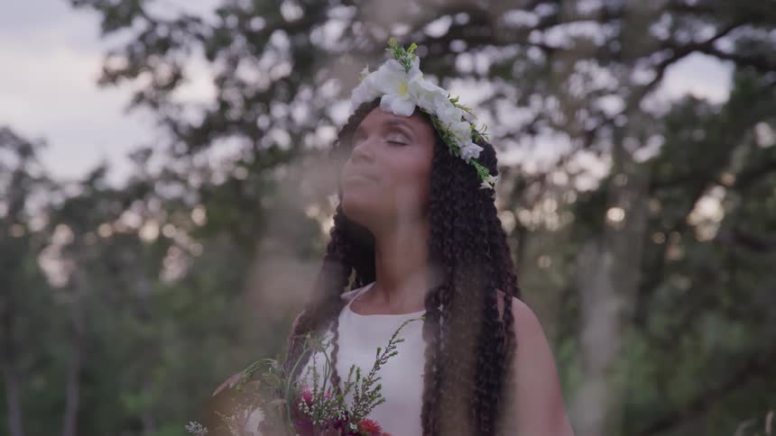 A woman holding a bouquet of flowers with flowers in her hair stands with her eyes closed